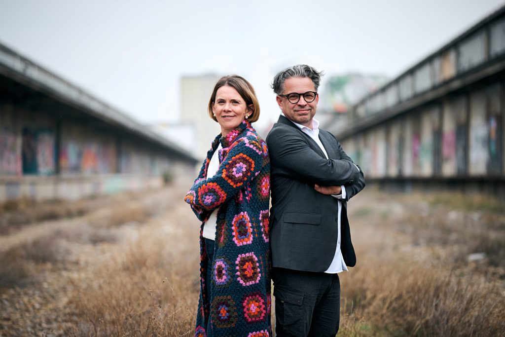 karin und hannes stehen Rücken an Rücken an einem Los Place und blicken in die Kamera